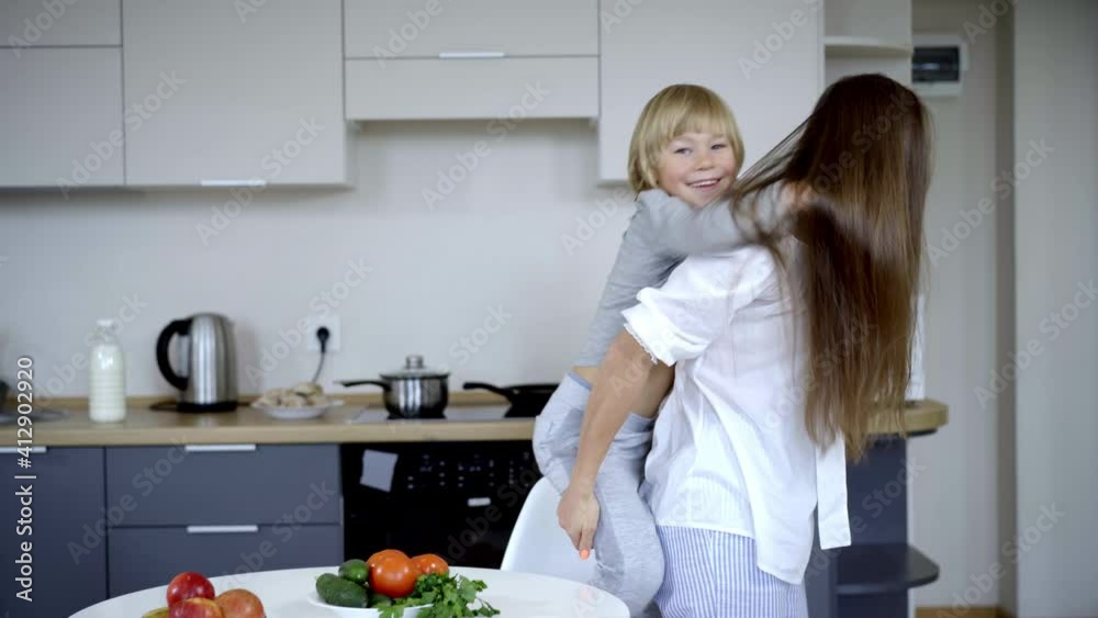 Cheerful Caucasian mother entering kitchen holding son on back and hugging kid. Portrait of happy positive Caucasian family enjoying morning at home. Domestic life concept. Cheerful Caucasian mother entering kitchen holding son on back and hugging kid. Portrait of happy positive Caucasian family enjoying morning at home. Domestic life concept.