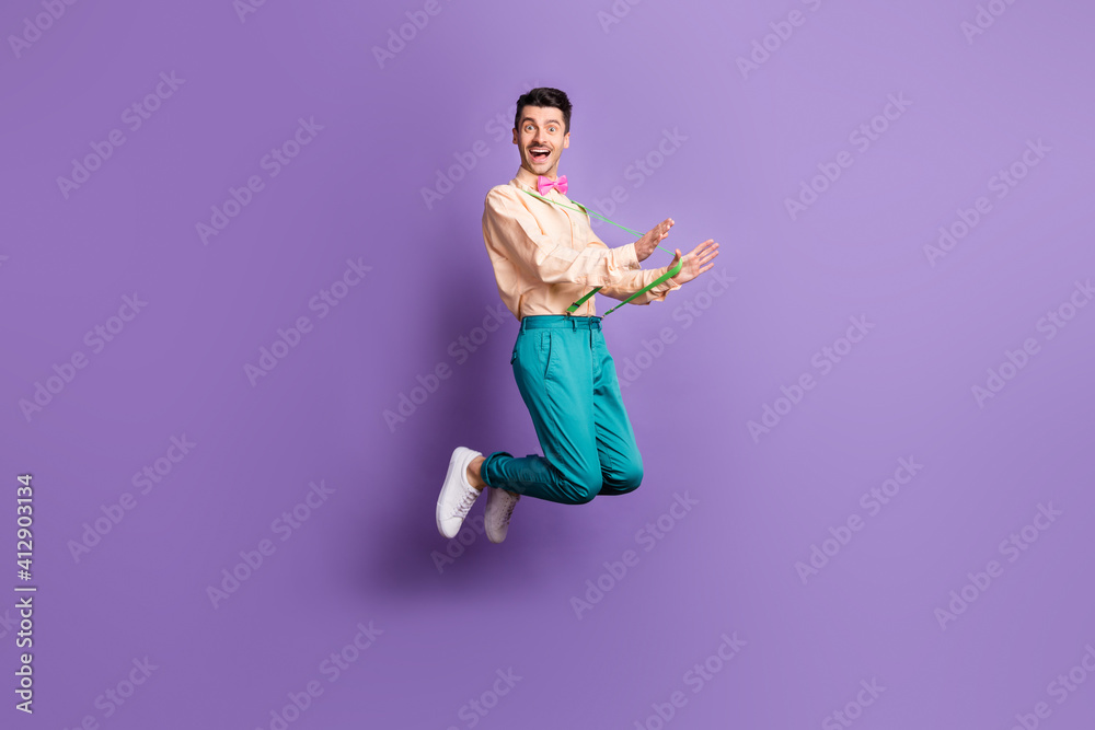 Full length profile side photo of happy crazy funky man pull suspenders jump up air isolated on violet color background Full length profile side photo of happy crazy funky man pull suspenders jump up air isolated on violet color background