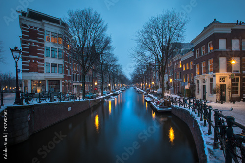 Winter snow view of Dutch canal and old houses in the historic city of Amsterdam during the blue hour, the Netherlands