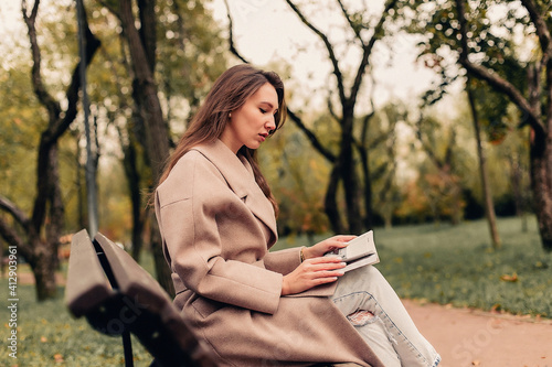 Bild auf Leinwand A Caucasian girl in a beige coat and blue jeans sits on a bench in a park in aut