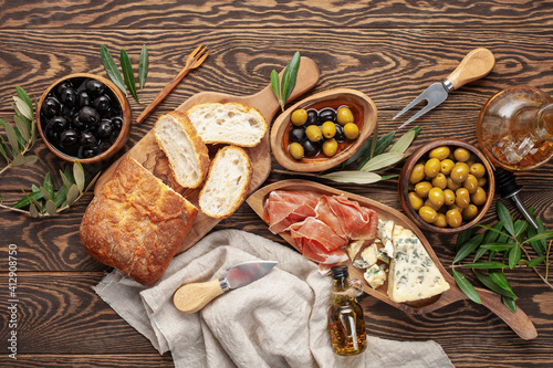 Black and green Olives, olive tree branch, oil, cheese Dorblu or Gorgonzola, prosciutto and ciabatta bread on dark wooden table. Top view. Flat lay