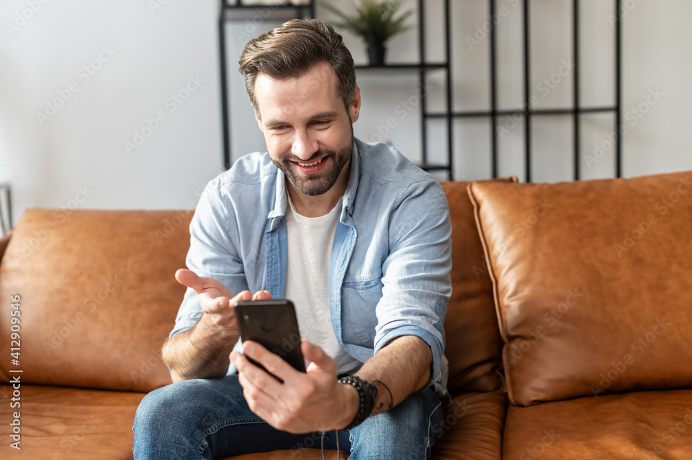 Smiling guy wearing casual outfit talks by video connect on the smartphone. Front view a young man sits on the modern sofa and using mobile app for virtual meeting, video conference Smiling guy wearing casual outfit talks by video connect on the smartphone. Front view a young man sits on the modern sofa and using mobile app for virtual meeting, video conference