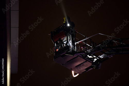 Firefighters rise on a mechanical sliding ladder to the epicenter of the fire. Fighting fire from bucket atop a fire truck. A fireman's crane in action, silhouette against a dark night sky.
