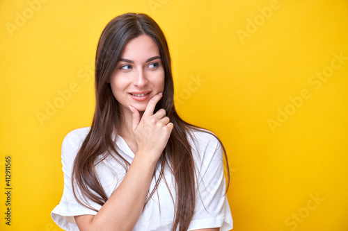 Portrait of brunette girl smiling, holding her chin and thinking, looks to the side at the copy space isolated on yellow background