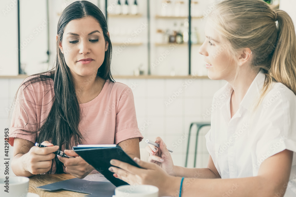 Young agent presenting content on tablet screen to female customer. Two women discussing documents and using device over cup of coffee. Consulting or meeting concept Young agent presenting content on tablet screen to female customer. Two women discussing documents and using device over cup of coffee. Consulting or meeting concept