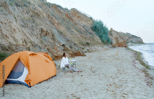 Summer beach camping. Mixed race woman sitting by orange tent in joyful mood enjoying beautiful sea view. Concept people and nature