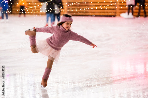 Little girl figure skater in a pink sweater is skating on winter evening on an outdoor ice rink lit by garlands photo