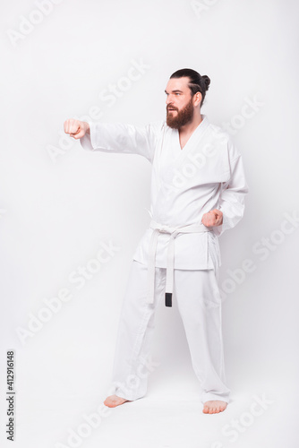 Photo of young bearded man in dobok uniform practicing taekwondo over white background.