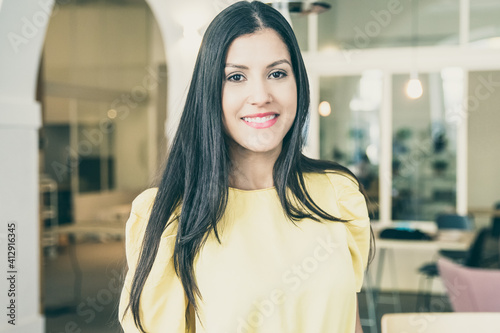 Happy gorgeous black haired woman wearing yellow shirt, standing in co-working space, posing, looking at camera and smiling. Female portrait concept