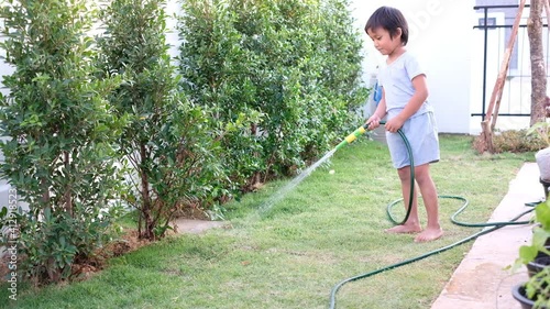 Funny Asian little boy dancing while watering plants in the garden. Cute child using garden hose in the backyard.