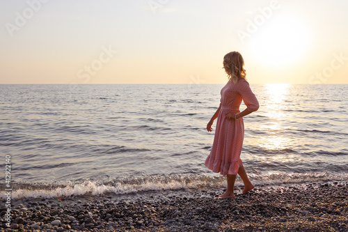 Young woman in pink dress slowly walks along the coastline of surf barefoot and looks at horizon, with copy space