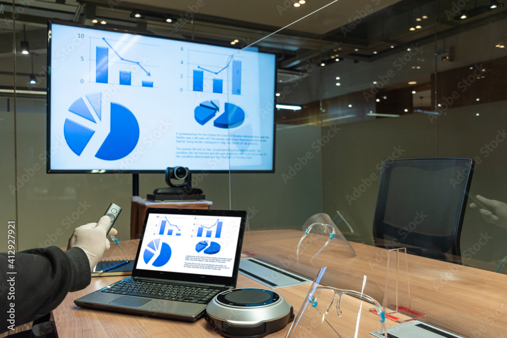 Naklejka premium Man wearing rubber glove with a remote control slide presentation with Presentation meeting on television and laptop in meeting room with clear acrylic sheet separates the center on table