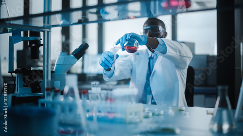 African American Male Scientist Wearing Protective Goggles Mixing Chemicals in a Test Tube in a Lab. Handsome Black Microbiologist Working in Modern Laboratory with Technological Equipment.