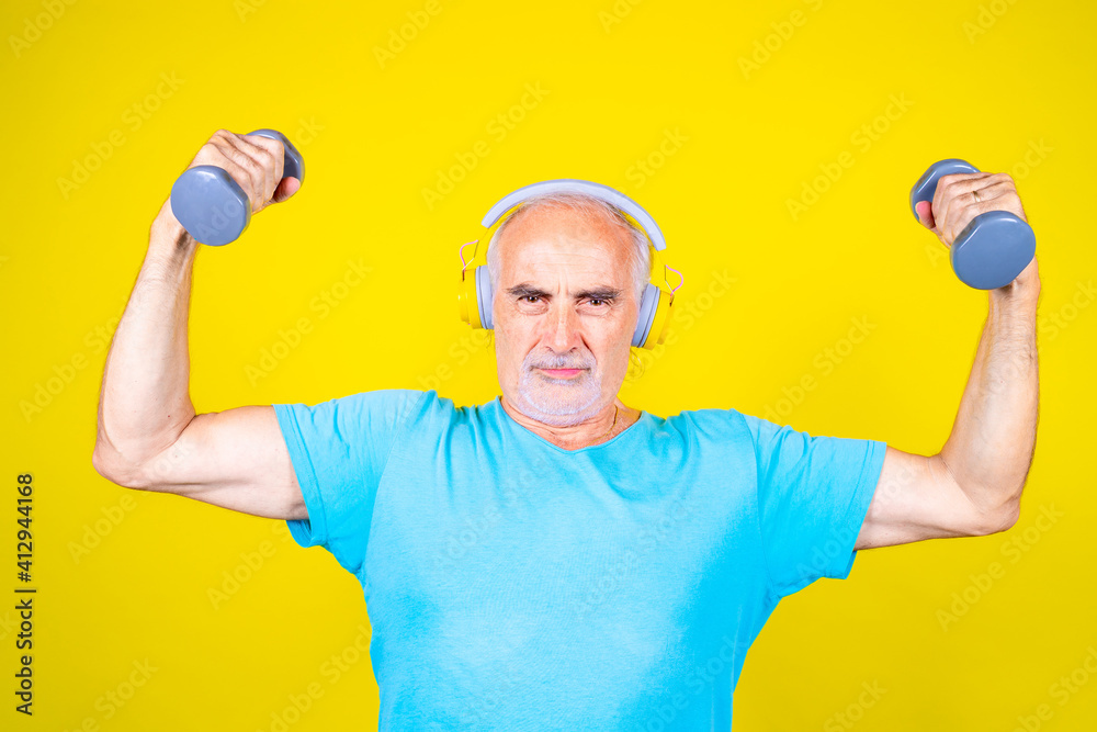 Elderly man training using dumbbells on yellow background smiling stretching using gym weight listening music Elderly man training using dumbbells on yellow background smiling stretching using gym weight listening music