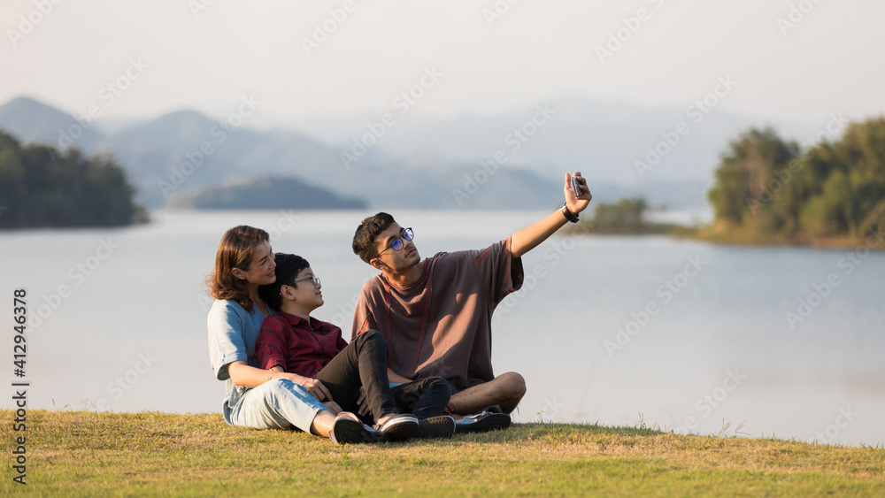 Asian family three members, mother and two young sons, sitting together beside huge lake with mountains and water in background. They using smartphone to take photos