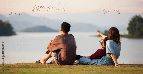 Mother and two sons sitting beside a big lake and see mountain view in the background, mom pointing finger to birds flying in sky. Idea for family tourist travels together