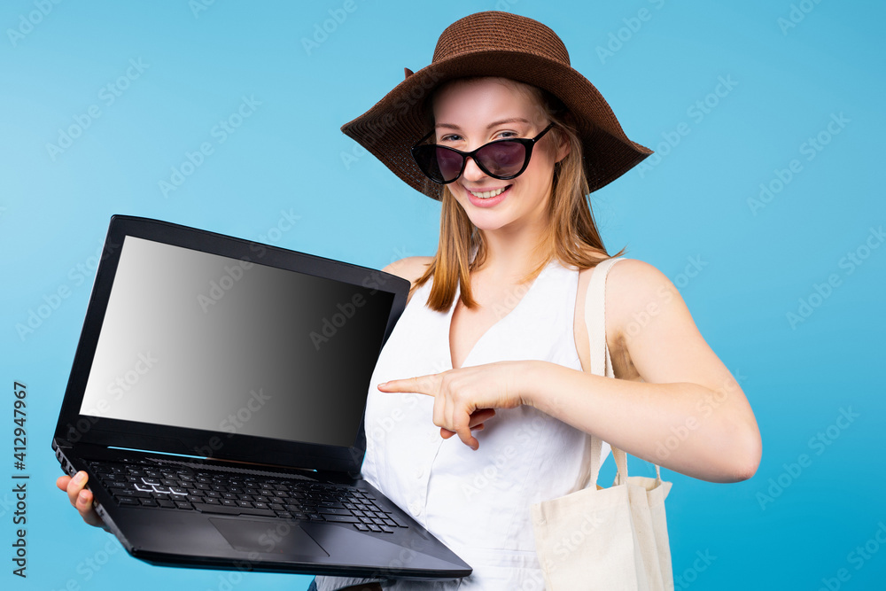 A beautiful blonde girl with sunglasses on her head and hat points to a laptop monitor. Blue background