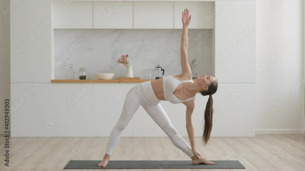 Young attractive multi ethnical woman wearing white sportswear, pants and top, doing stretching yoga exercises on fitness mat in living room, healthcare and exercise mental therapy at home concept