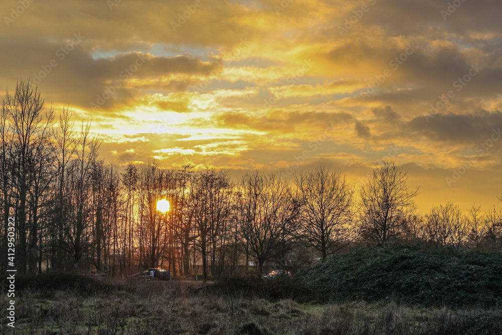 Fototapeta premium Dutch countryside with a beautiful sunset with the sun between the branches of the bare trees and the clouds on a cloudy afternoon, magical moment in Dal van de Roodebeek in South Limburg, Netherlands