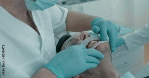 During dentist`s appointment male Arabian patient is having his gum examined by dentist. Patient is having cheek retractor and protective eyeswear on him.