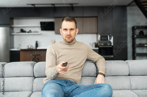 Handsome bearded young man holding tv remote control, switch channels, watching evening TV shows, spends leisure time on the couch at home in front of television set © Vadim Pastuh