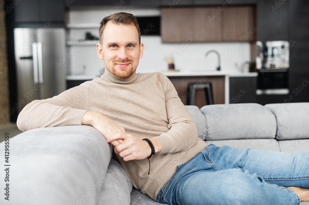 Portrait of handsome young man having rest in modern living room at home, sitting casually on sofa, looks at the camera, feeling relaxed. Lifestyle and leisure