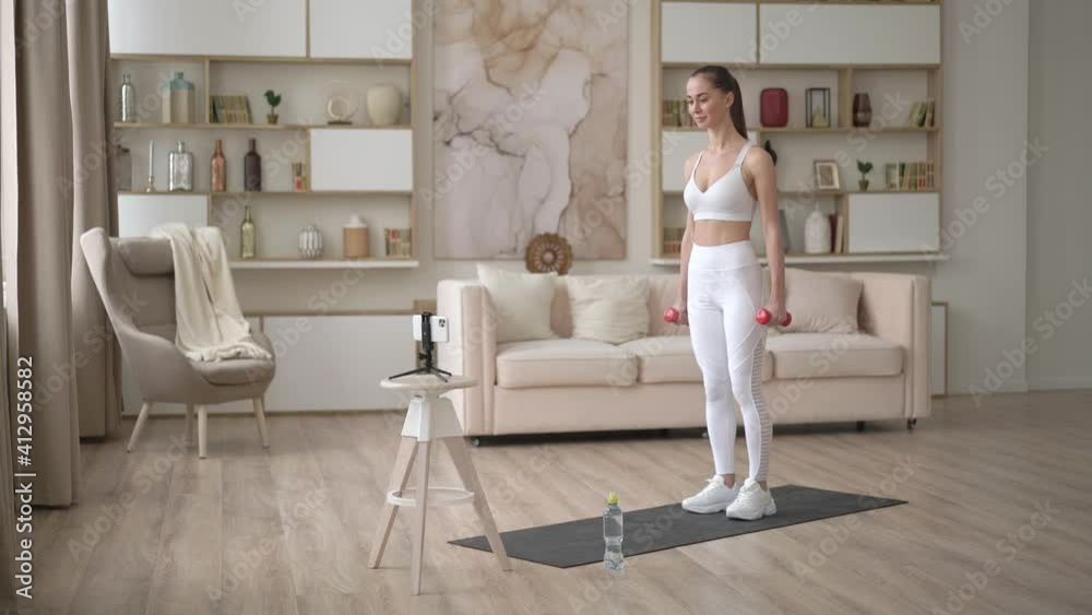 Sporty young woman doing online workout exercise with dumbbells. Active girl enjoy sport fitness training on smartphone at home, stretching on yoga mat watching video. Sport and recreation concept. Sporty young woman doing online workout exercise with dumbbells. Active girl enjoy sport fitness training on smartphone at home, stretching on yoga mat watching video. Sport and recreation concept.