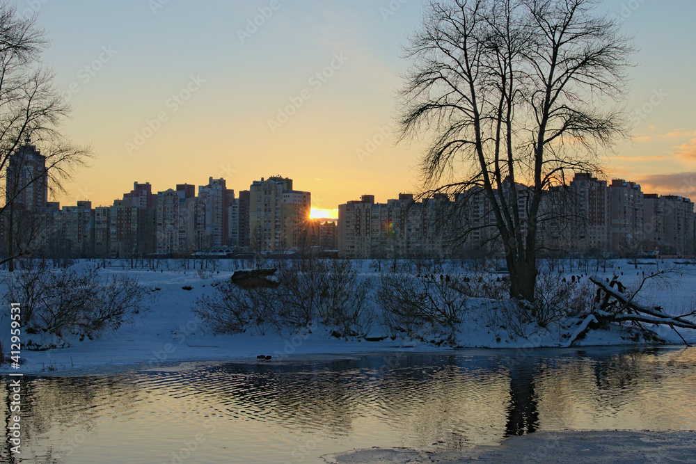 Obraz premium Picturesque winter landscape of frozen Dnieper River during sunset. Buildings of The Obolon neighborhood in the background. Bare tree reflected in the water. The sun is hiding behind the house. Kyiv