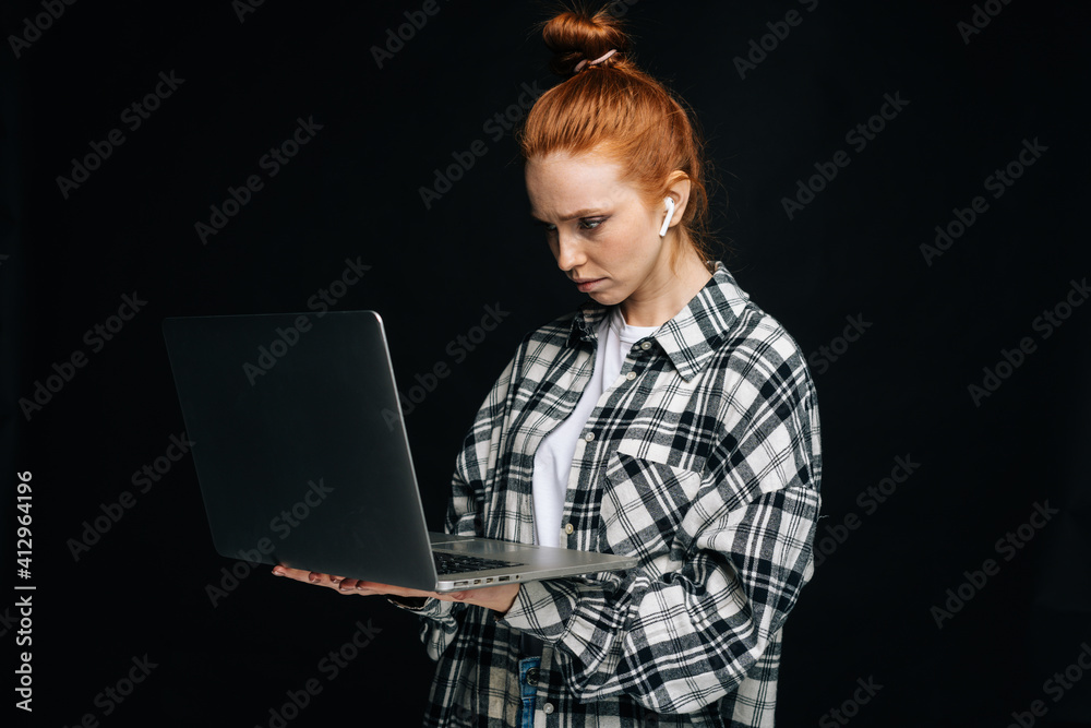 Focused cute young woman holding laptop computer and working typing on isolated black background. Pretty redhead lady model emotionally showing facial expressions in studio, copy space.