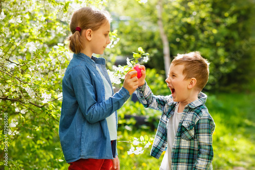 A boy and a girl are having fun on a holiday and breaking Easter eggs in the garden against the background of cherry blossoms.
