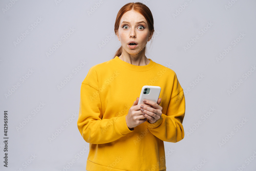 Portrait of shocked cute young woman holding mobile phone and looking at camera on isolated white background. Pretty redhead lady model emotionally showing facial expressions in studio, copy space. Portrait of shocked cute young woman holding mobile phone and looking at camera on isolated white background. Pretty redhead lady model emotionally showing facial expressions in studio, copy space.