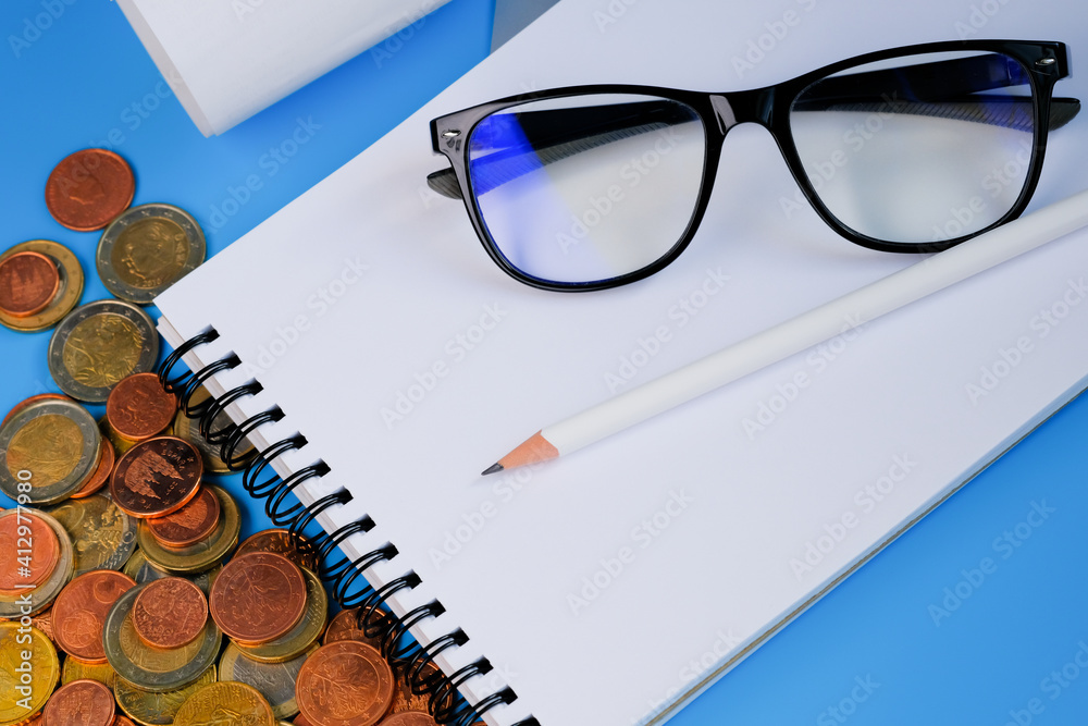 Finance planning and economy concept. Notebook, pencil, glasses and coins on blue background