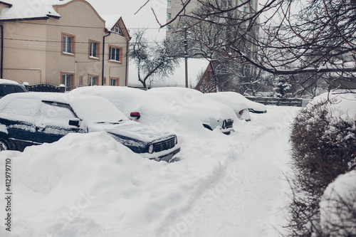 Parked cars covered with snow during snowfall in city. Automobiles stuck in heaps after winter blizzard