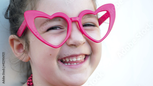 Close up. Cute brown eyes kid in red heart glasses smiles showing tongue at camera, white background. Valentines Day © IULIIA
