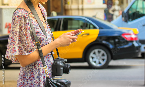 A girl in a floral summer sundress with a camera is typing a text message or looking for information on smartphone. Citizens on the street. The black and yellow cab in the background.