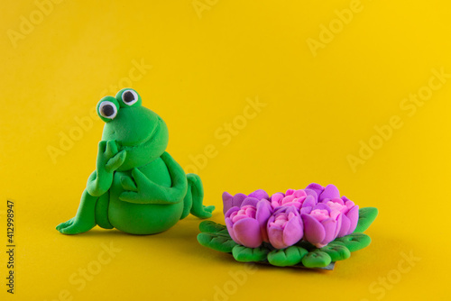 Dreamy frog figure made of light clay modeline sitting and holding head with hand near flower bouquet on yellow background © virginna
