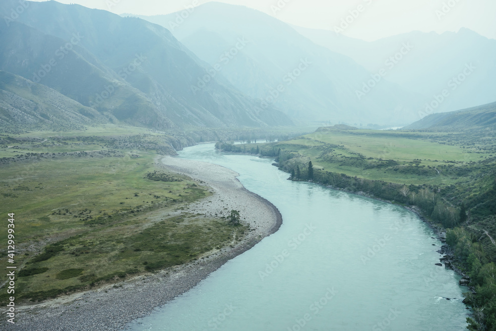Beautiful misty mountain landscape with wide mountain river. Dark green gloomy scenery with big mountain river in mist. Dark atmospheric view to great river among big mountains in rainy weather.