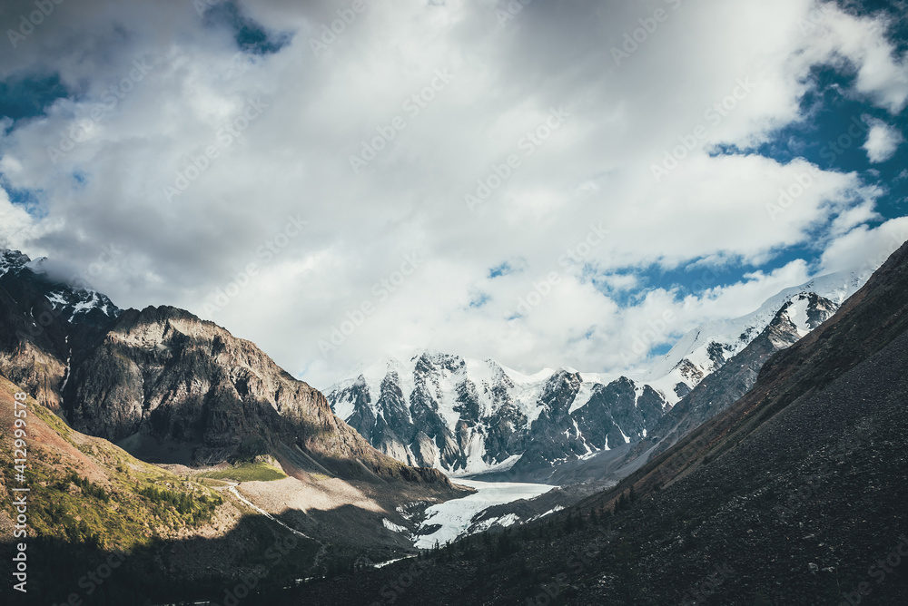 Beautiful mountain landscape with glacier tongue and great snowy mountains in sunlight in cloudy sky. Scenic alpine landscape with glacial tongue and high mountains with glaciers in low clouds.