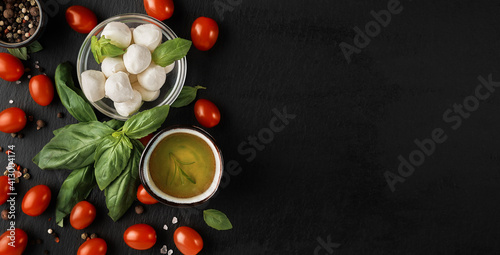 Mozzarella and cherry tomatoes with basil leaves, salt and pepper, layout on a black stone board. Ingredients for making Caprese salad. Top view with copy space for text