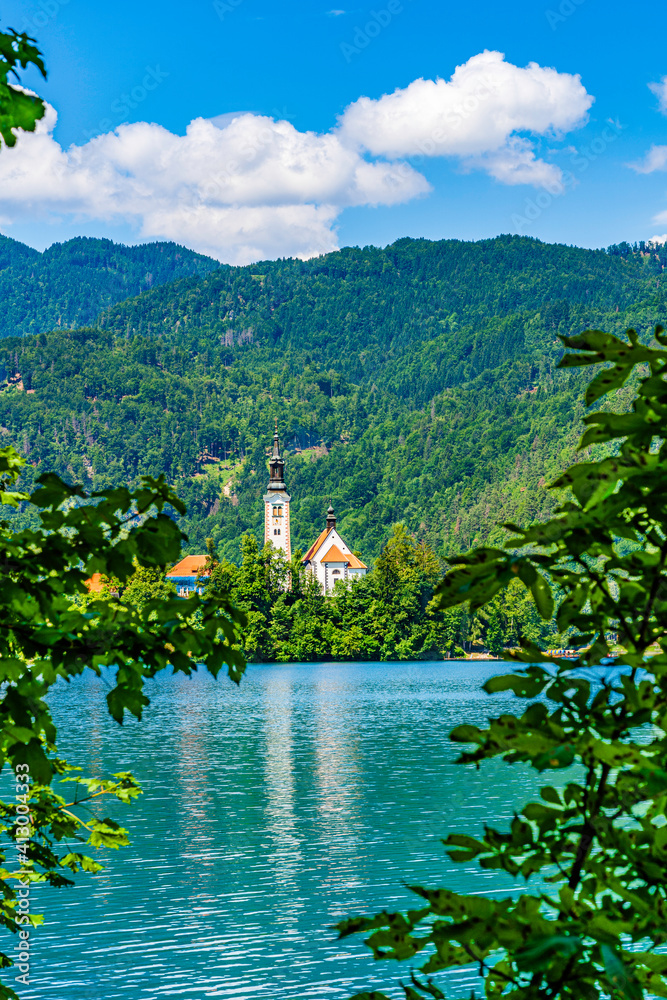 Vertical landscape of Lake Bled in Slovenia with the small church island and Alps mountains in the background seen thrugh tree branches