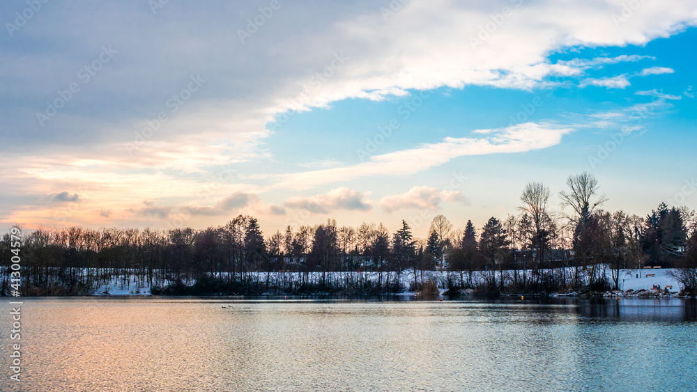 Winter ladnscape at lake at sunset, reflections in water mirror, sunlit blue sky clouds in backgorund