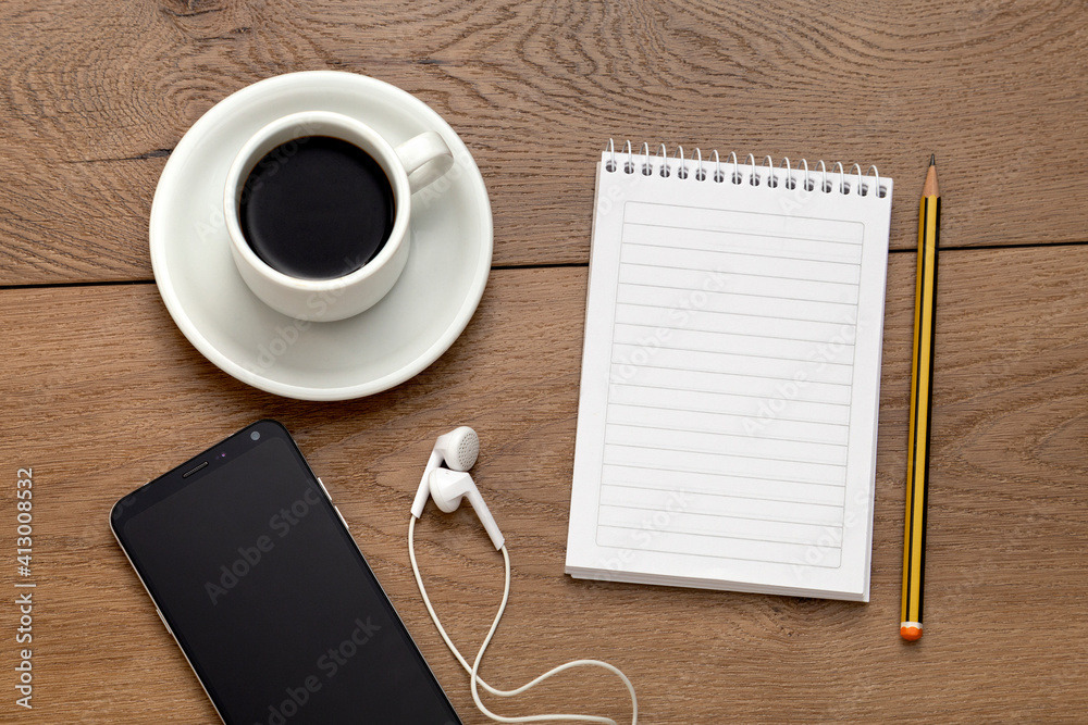 Blank notebook next to a pencil, a cup of coffee and phone with headphones. All over a wooden table. Special for mockup.