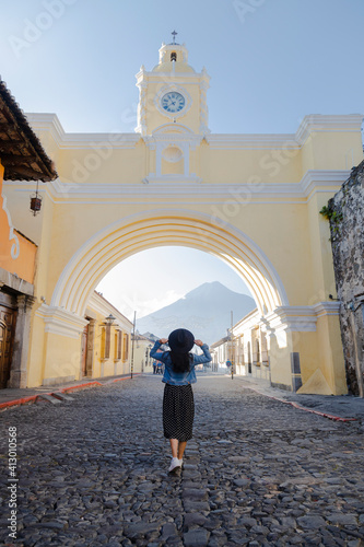 Young Hispanic woman walking under the Arch of Santa Catalina in Antigua Guatemala - tourist in important avenue of colonial Guatemala city with the Water volcano background