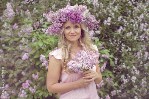 Blonde woman enjoying in a spring garden with blooming lilacs with bouquet, lilac flowers hair style.  Spring Concept, Spring blossom, Positive human emotions and feelings. 