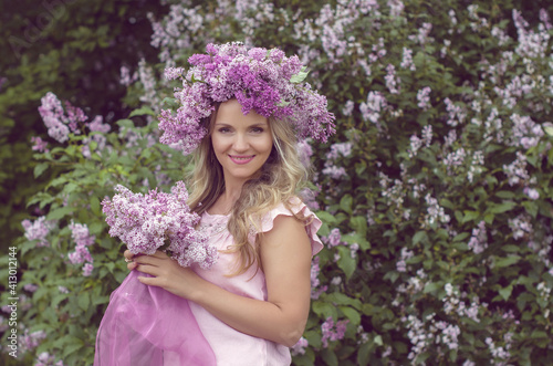 Blonde woman enjoying in a spring garden with blooming lilacs with bouquet, lilac flowers hair style. Spring Concept, Spring blossom, Positive human emotions and feelings. 