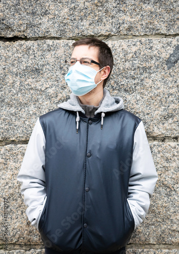 Vertical shot of a European tourist in glasses and facial mask standing near a wall photo