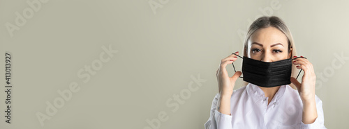 young woman blonde doctor cosmetologist in a white coat holding a black disposable mask to dress on her face, business banner on a light background