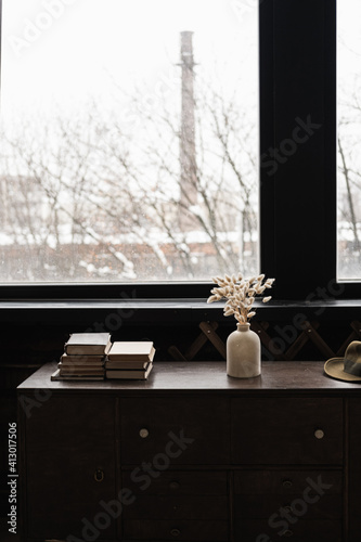 Fototapeta Naklejka Na Ścianę i Meble -  Rabbit bunny tail grass bouquet, books stack on solid wooden table against window. Aesthetic dark home interior design concept. Beautiful floral foliage composition.