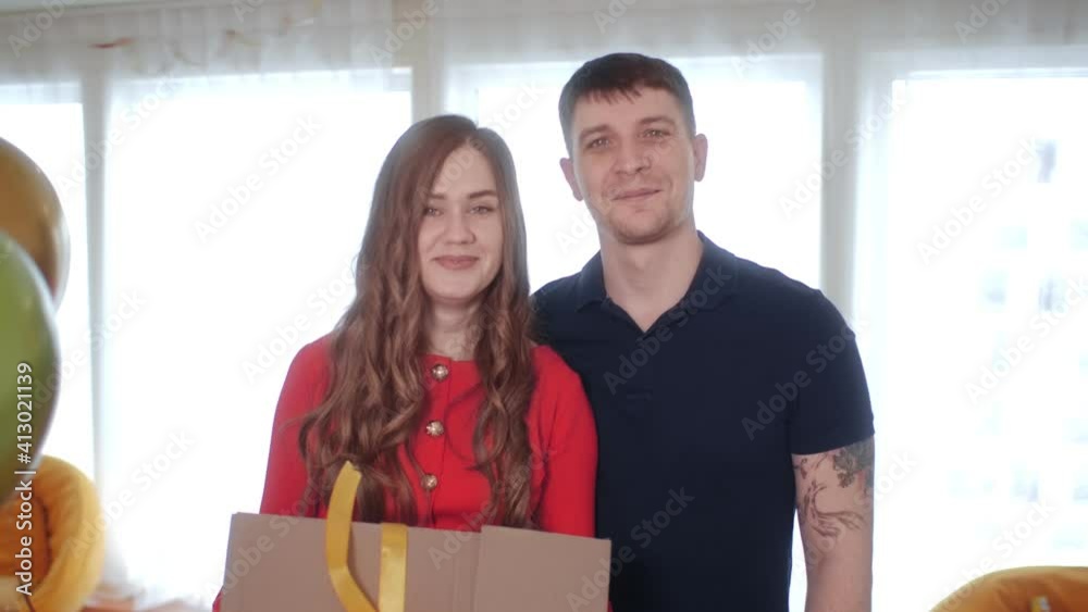 Beautiful young couple with a big gift box standing in a room with flying balloons and smiling to the camera. Balloons fly in front of the camera. Beautiful young couple with a big gift box standing in a room with flying balloons and smiling to the camera. Balloons fly in front of the camera.