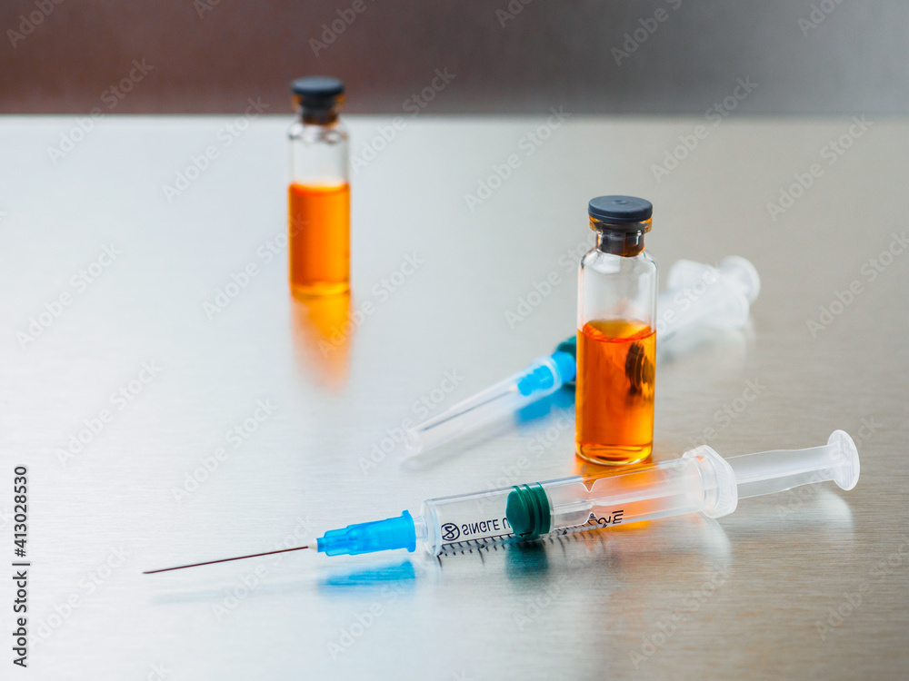Selective focus. Close-up shot of injection syringes and ampoules with orange coronavirus vaccine on a silver aluminum background. Covid-19 pandemic medical concept. Copy space. Selective focus. Close-up shot of injection syringes and ampoules with orange coronavirus vaccine on a silver aluminum background. Covid-19 pandemic medical concept. Copy space.